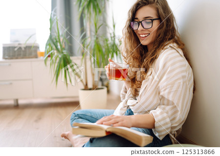 A young curly woman reads a book and drinks coffee or tea while sitting on floor in the living room. A young curly woman reads a book and drinks coffee or tea while sitting on floor in the living room. 125313866