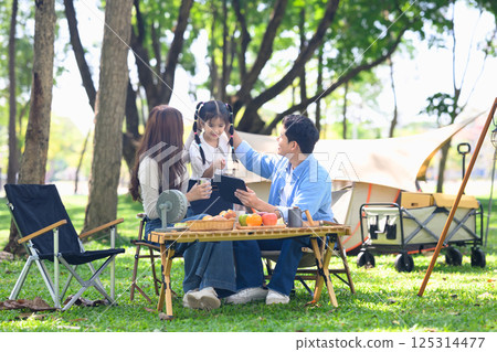 Happy family enjoying a picnic together at campsite with foods and fresh fruit on a sunny day Happy family enjoying a picnic together at campsite with foods and fresh fruit on a sunny day 125314477