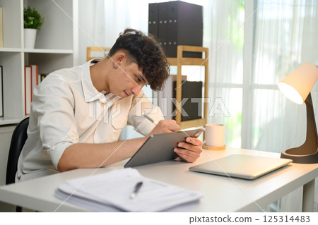 Focused young man writing with stylus on digital tablet working at a home office desk. 125314483
