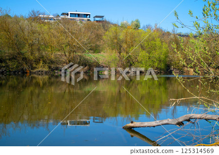 Modern private house over the river. The modern house stands on a hill above the river. The calm flow of the river surrounded by spring nature. Modern private house over the river. The modern house stands on a hill above the river. The calm flow of the river surrounded by spring nature. 125314569