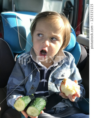 Child in car holding cucumber and apple and looking angry Child in car holding cucumber and apple and looking angry 125315115