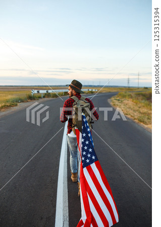 4th of July. American Flag. Traveler with the flag of America. The man in a hat, a backpack, a shirt and jeans. 4th of July. American Flag. Traveler with the flag of America. The man in a hat, a backpack, a shirt and jeans. 125315394