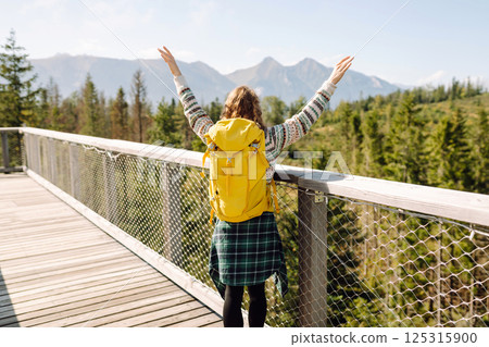 Happy woman with yellow backpack on wooden pedestrian bridge enjoying mountain scenery. Happy woman with yellow backpack on wooden pedestrian bridge enjoying mountain scenery. 125315900