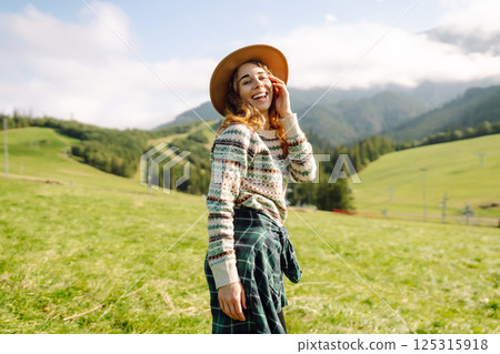 Stylish female traveler in hat has fun on top of hill, enjoys mountain landscape. Active lifestyle. 125315918