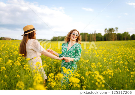Two young women in beautiful dresses walk in a field with yellow blooming rapeseed. Concept of fun. Two young women in beautiful dresses walk in a field with yellow blooming rapeseed. Concept of fun. 125317206