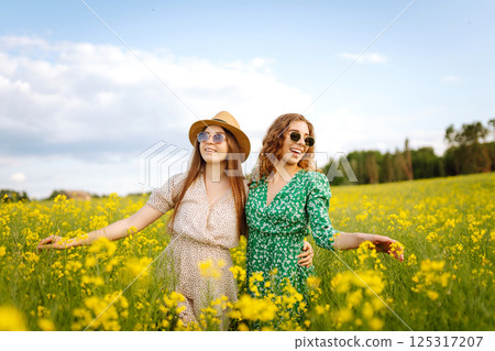 Two young women in beautiful dresses walk in a field with yellow blooming rapeseed. Concept of fun. 125317207