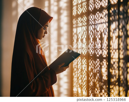 Woman in a hijab praying at home, holding a misbaha, with soft light through patterned windows 125318966