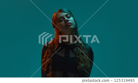 neon portrait of young woman in sheer black blouse tilting head gently in dramatic teal-orange light against gradient background. neon portrait of young woman in sheer black blouse tilting head gently in dramatic teal-orange light against gradient background. 125319493