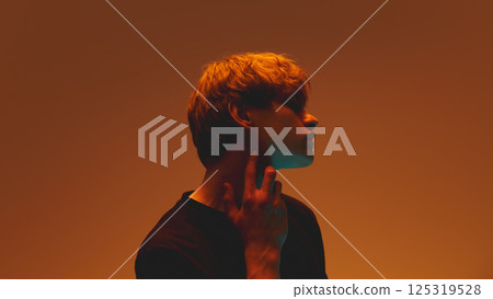 Portrait of young man with tense expression, pressing fingers to neck, teal glow on face highlight appearance against warm background. 125319528