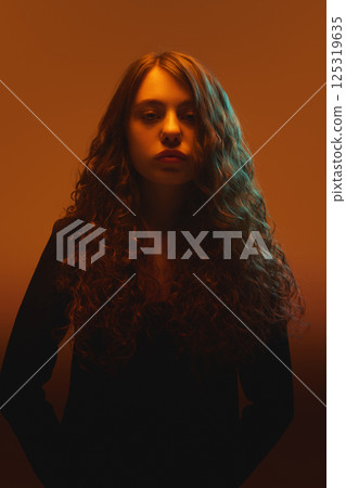 Portrait of beautiful woman with loose curls facing camera with neutral expression in warm neon light against gradient background. Portrait of beautiful woman with loose curls facing camera with neutral expression in warm neon light against gradient background. 125319635