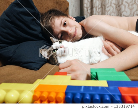A cheerful young boy hugging his white and gray cat while lying on the couch, portraying companionship and love. happy boy cuddles with his friend the cat A cheerful young boy hugging his white and gray cat while lying on the couch, portraying companionship and love. happy boy cuddles with his friend the cat 125319714
