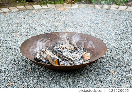 A close-up shot of burning firewood in a shallow, rusted steel fire pit placed on a gravel surface. Light smoke rises into the air while small flames dance around the blackened logs.  125320095