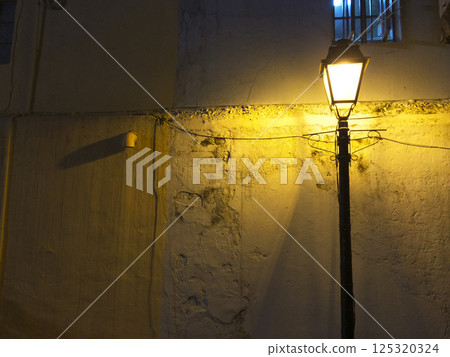 夜晚，後巷黑路燈發出溫暖的橘色燈光，牆面剝落，窗戶上裝有鐵條 125320324