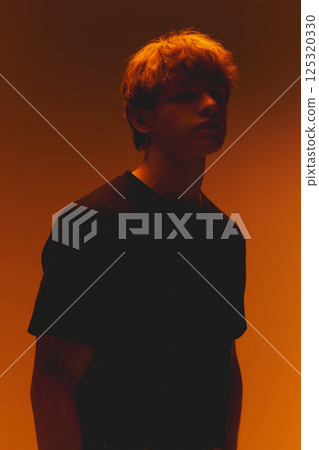 Young man in shadows, face partially hidden, in warm dramatic neon lighting against warm orange gradient background. Concept of beauty Young man in shadows, face partially hidden, in warm dramatic neon lighting against warm orange gradient background. Concept of beauty 125320330