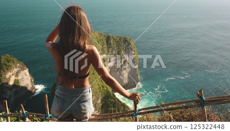 Young woman is taking in the beauty of the turquoise ocean and the dramatic cliffs of kelingking beach from a scenic viewpoint Young woman is taking in the beauty of the turquoise ocean and the dramatic cliffs of kelingking beach from a scenic viewpoint 125322448