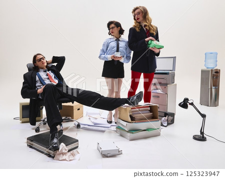 Man leaning back with hands behind head while two women in formal outfits stand holding cup and phone in cluttered workspace. Man leaning back with hands behind head while two women in formal outfits stand holding cup and phone in cluttered workspace. 125323947