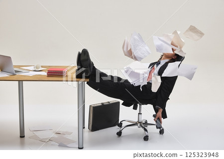 Man sitting in chair with legs on desk, surrounded by flying papers in chaotic office mess, overwhelmed by work. Dynamic photo. 125323948
