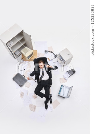 Overhead ew of man in black suit reclining on chair with papers and phone in hand surrounded by office mess and outdated tech. 125323955