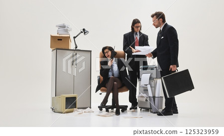 Three office workers gathered around printer, woman yelling on phone while others present documents in messy white studio scene. 125323959
