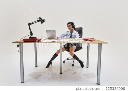 Young woman in shirt and tie working with laptop and documents on messy table, wearing headphones and focused expression in office setup. 125324008