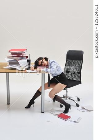 Young woman resting head on documents, leaning on desk with sleepy expression in messy office setup against white studio background. 125324011