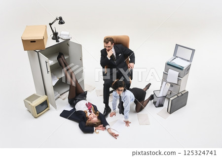 Boss seated at center while two female workers lie exhausted on floor in chaotic office scene filled with paper and clutter. Boss seated at center while two female workers lie exhausted on floor in chaotic office scene filled with paper and clutter. 125324741