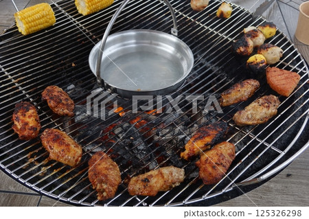 close up of barbecue over an open flame with a grill, chicken wings and corns are sizzling over glowing coals with a hot pot in the middle close up of barbecue over an open flame with a grill, chicken wings and corns are sizzling over glowing coals with a hot pot in the middle 125326298