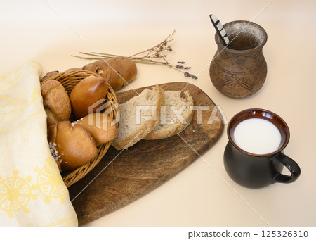 A cup of milk and a basket of buns and rolls, towel, dry flowers and a clay pot with a spoon 125326310