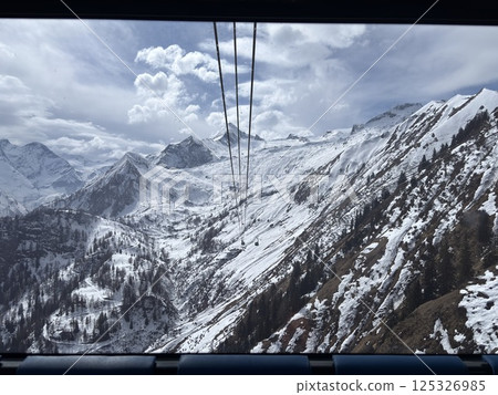 View from the cab of a cable car in the Alps, on a glacier in spring. Partially descended avalanches, partially peaks in the snow. Sun and clouds, high cable car. 125326985