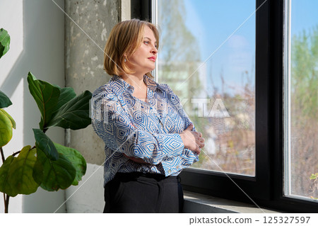 Portrait of beautiful middle-aged woman with crossed arms looking out window Portrait of beautiful middle-aged woman with crossed arms looking out window 125327597