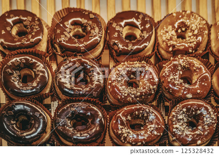 Rows of chocolate glazed donuts with toppings displayed in a pastry shop. Sweet temptation scene, dessert cravings concept, food background 125328432