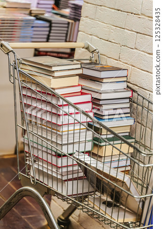 Metal shopping cart overflowing with hardcover books in bookstore aisle. Books in cart symbolize curiosity, discovery and endless learning Metal shopping cart overflowing with hardcover books in bookstore aisle. Books in cart symbolize curiosity, discovery and endless learning 125328435