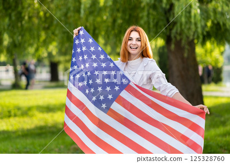 Portrait of happy young red haired woman holding USA national flag in her hands standing outdoors in summer park. Positive girl celebrating United States independence day. 125328760