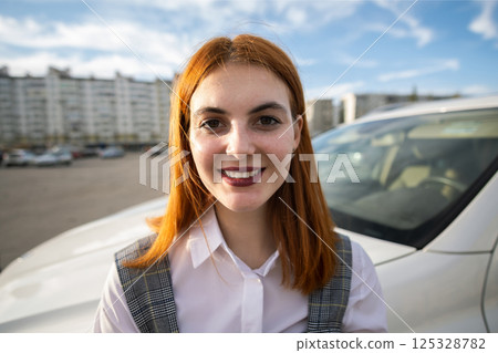 Portrait of a fashionable teenage girl with red hair wearing casual dress standing outside in front of big new expensive car on warm summer day. 125328782