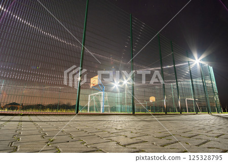 Outdoors mini football and basketball court with ball gate and basket surrounded with high protective fence brightly illuminated with spotlight lamps at night. Outdoors mini football and basketball court with ball gate and basket surrounded with high protective fence brightly illuminated with spotlight lamps at night. 125328795