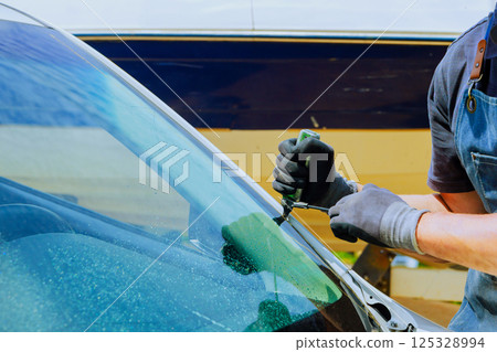 Technician replaces damaged windshield using special tools with precision while working at an auto service center. 125328994