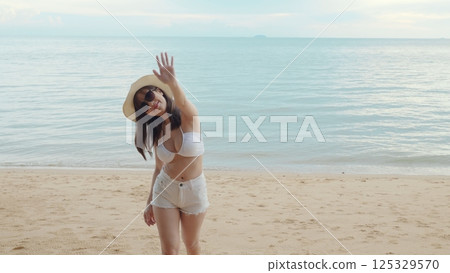 Beautiful Asian woman wear sungrass and hat turning back running to tropical paradise beach, Happy female in bikini having fun with hand to camera inviting you come with me 125329570