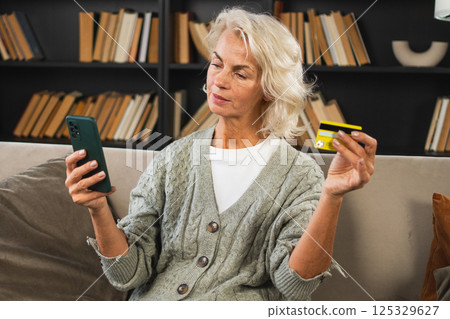 Happy middle aged woman shopping online with smartphone paying with credit card. Mature lady buying on Internet enter credit card details at home indoors. Online shopping delivery service ecommerce Happy middle aged woman shopping online with smartphone paying with credit card. Mature lady buying on Internet enter credit card details at home indoors. Online shopping delivery service ecommerce 125329627