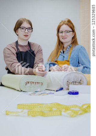 Two female seamstresses and fashion designers are standing at table with roll of fabric. Two female seamstresses and fashion designers are standing at table with roll of fabric. 125329685