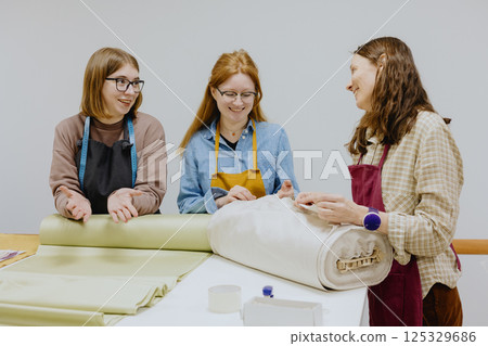 Three female seamstresses fashion designers stand at a table with roll of fabric and negotiate. Three female seamstresses fashion designers stand at a table with roll of fabric and negotiate. 125329686