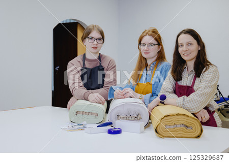 Three female seamstresses and fashion designers are standing at a table with a roll of fabric Three female seamstresses and fashion designers are standing at a table with a roll of fabric 125329687