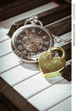 A retro pocket watch or Antique pocket watch with Golden heart shaped love padlock on Piano keys. 125331315