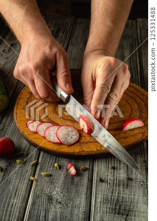 Hands skillfully slice radishes into thin rounds using a sharp knife on a rustic wooden cutting board, highlighting culinary precision and fresh ingredients in a cozy kitchen atmosphere 125331488