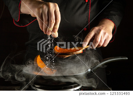 A chef is sprinkling seasoning over sweet potato slices in a pan, surrounded by steam. The moment captures the skill and concentration involved in culinary preparation 125331493