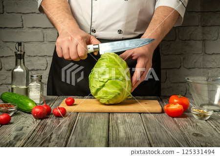 A chef in a white uniform skillfully cuts a head of cabbage on a wooden cutting board surrounded by fresh vegetables like tomatoes, cucumbers, and radishes 125331494