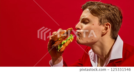 Side view humorous portrait of handsome young man in suit kissing delicious chicken burger against bold red background. 125331546