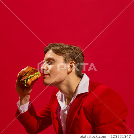 Satisfied young man in red suit holding delicious burger and smelling it with closed eyes against bold red background. Satisfied young man in red suit holding delicious burger and smelling it with closed eyes against bold red background. 125331547