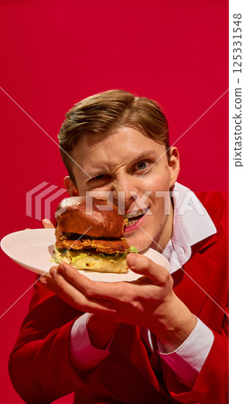 Portrait of emotional handsome young man in red suit holding plate with burger and biting small piece against bold red background 125331548