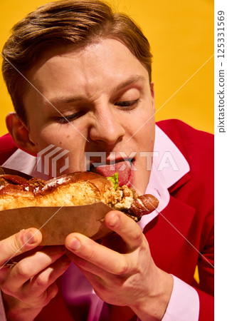 Close-up portrait of young man licking tip of hot dog with exaggerated expression and playful hunger against bright yellow background. 125331569