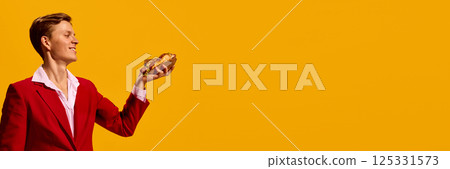 Smiling handsome man in red suit holding delicious hot dog against bold yellow background. Smiling handsome man in red suit holding delicious hot dog against bold yellow background. 125331573